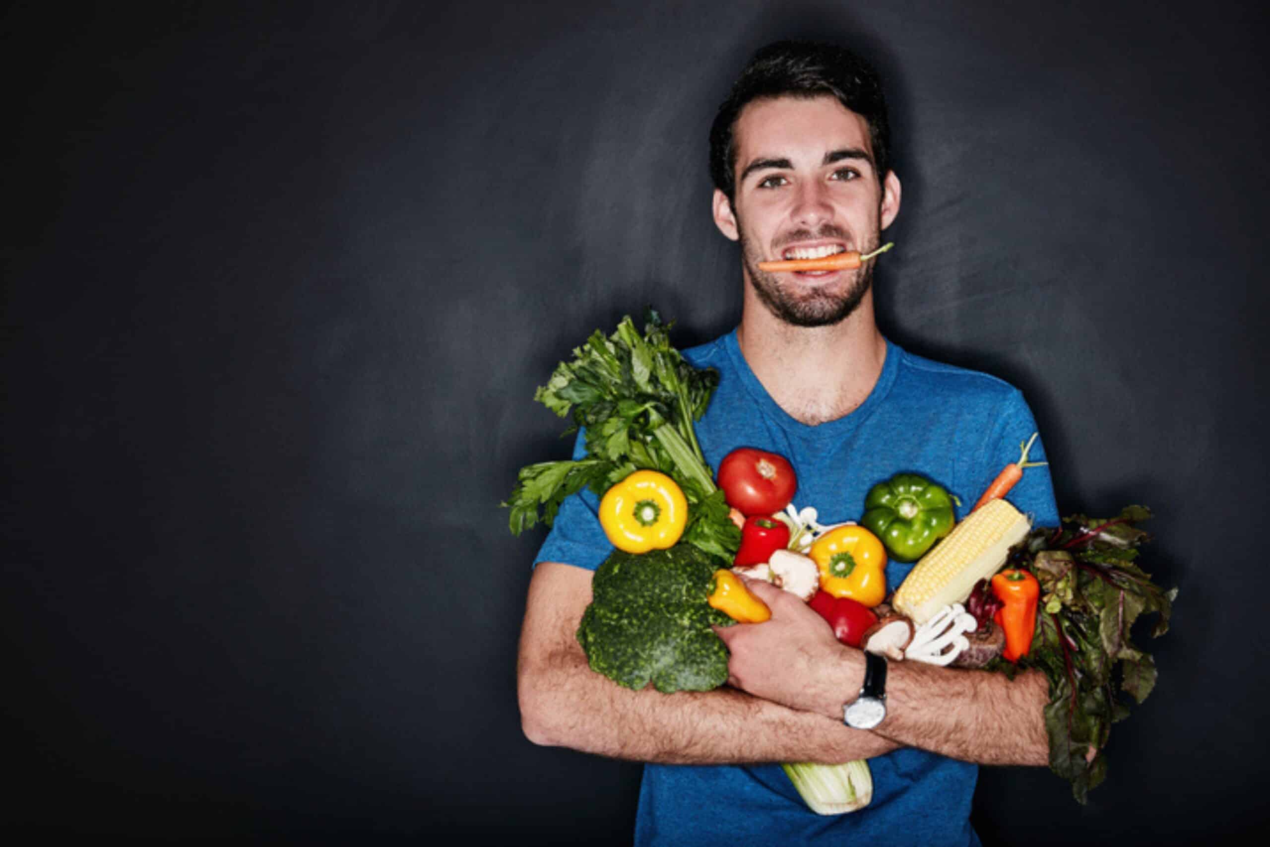 Smiling man holding assorted fresh vegetables with a carrot in his mouth